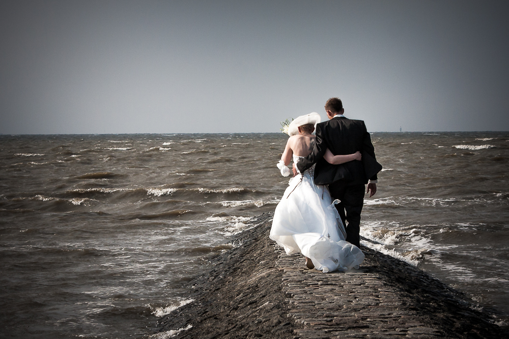 Braut und Bräutigam in Hochzeitskleidung gehen Hand in Hand entlang einer küstennahen Hochzeitslocation.