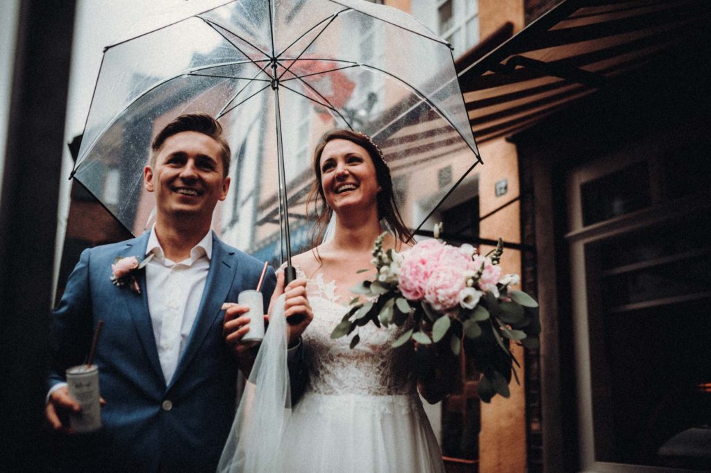 Fröhliche Braut und Bräutigam unter einem Regenschirm bei der Hochzeit, Braut trägt Blumenstrauß und Beide lächeln glücklich.