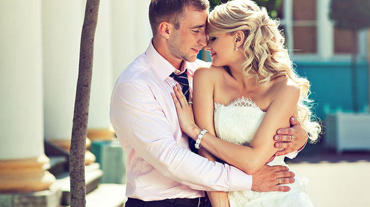 Braut und Bräutigam in Hochzeitskleidung umarmen sich vor der Hochzeitslocation, Hochzeitsfotografie bei Sonnenschein.