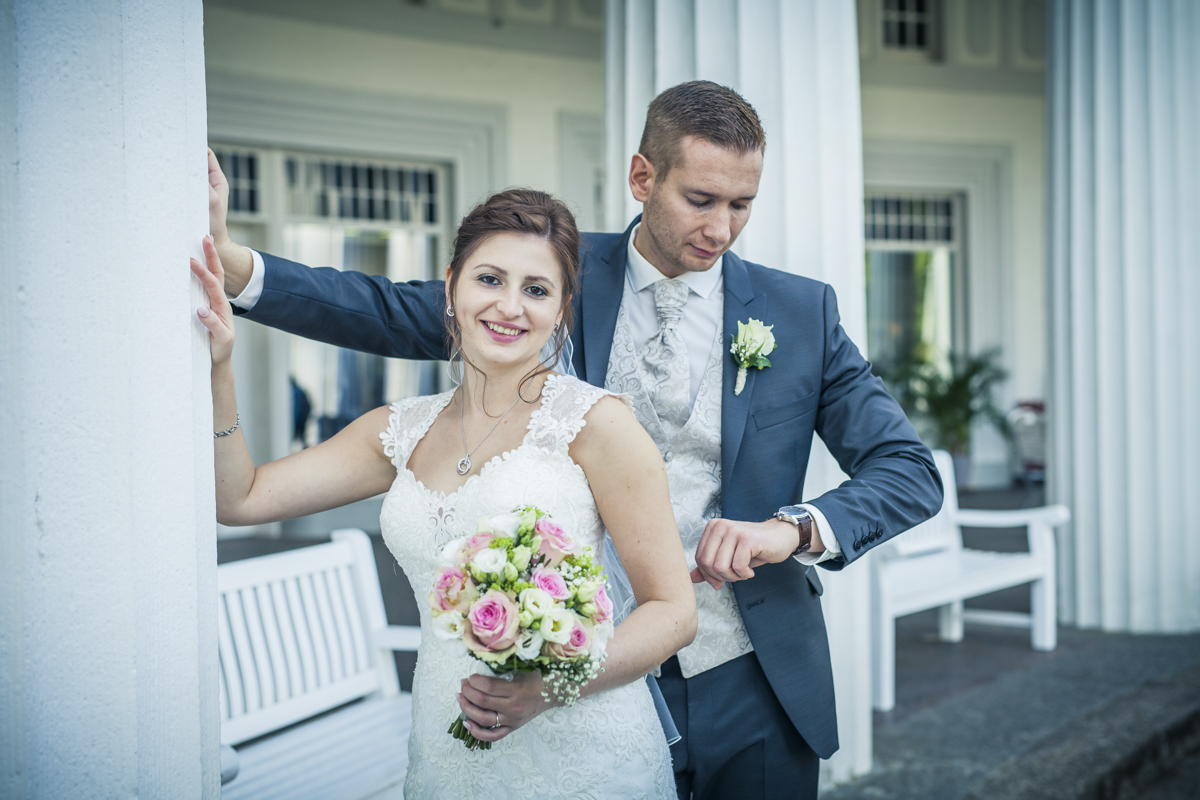 Braut im Hochzeitskleid mit Brautstrauß, Bräutigam kontrolliert Uhr, elegante Hochzeitsdekoration im Hintergrund