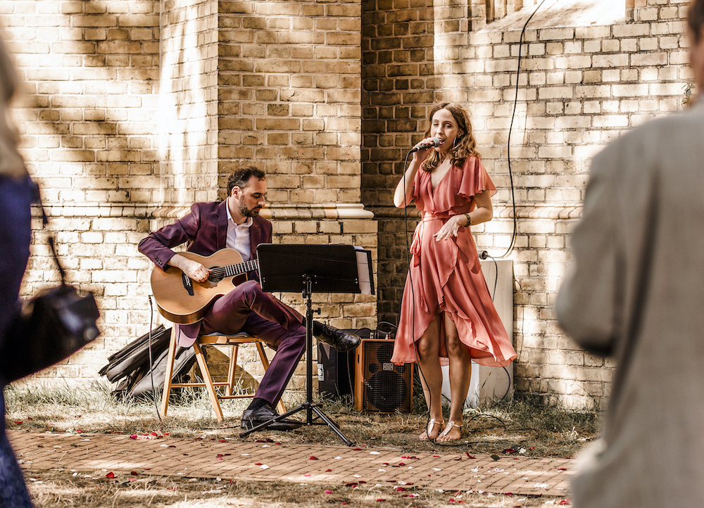 Musiker und Sängerin spielen Live-Musik bei einer Hochzeit im Freien, umgeben von Hochzeitsgästen und Dekoration.