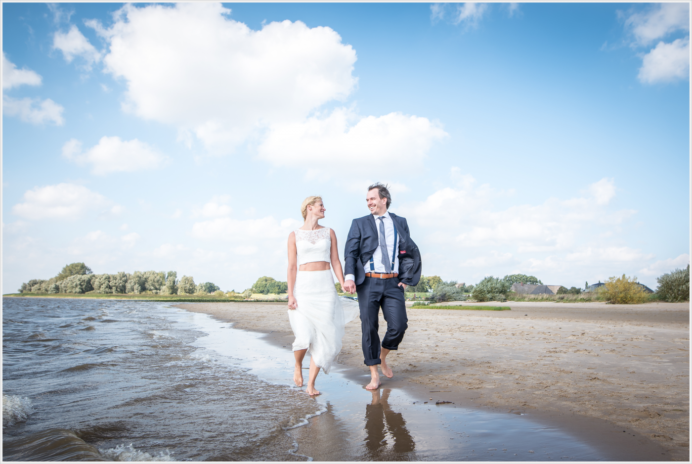 Braut und Bräutigam barfuß am Strand, sie trägt ein Hochzeitskleid, er einen Anzug, unter blauem Himmel am Meer.