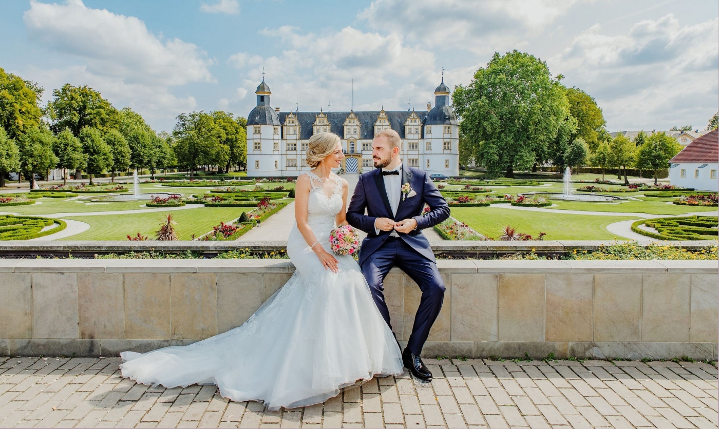 Braut und Bräutigam sitzen vor idyllischem Schlossgarten, sie hält einen Blumenstrauß, beide in Hochzeitskleid und -anzug.