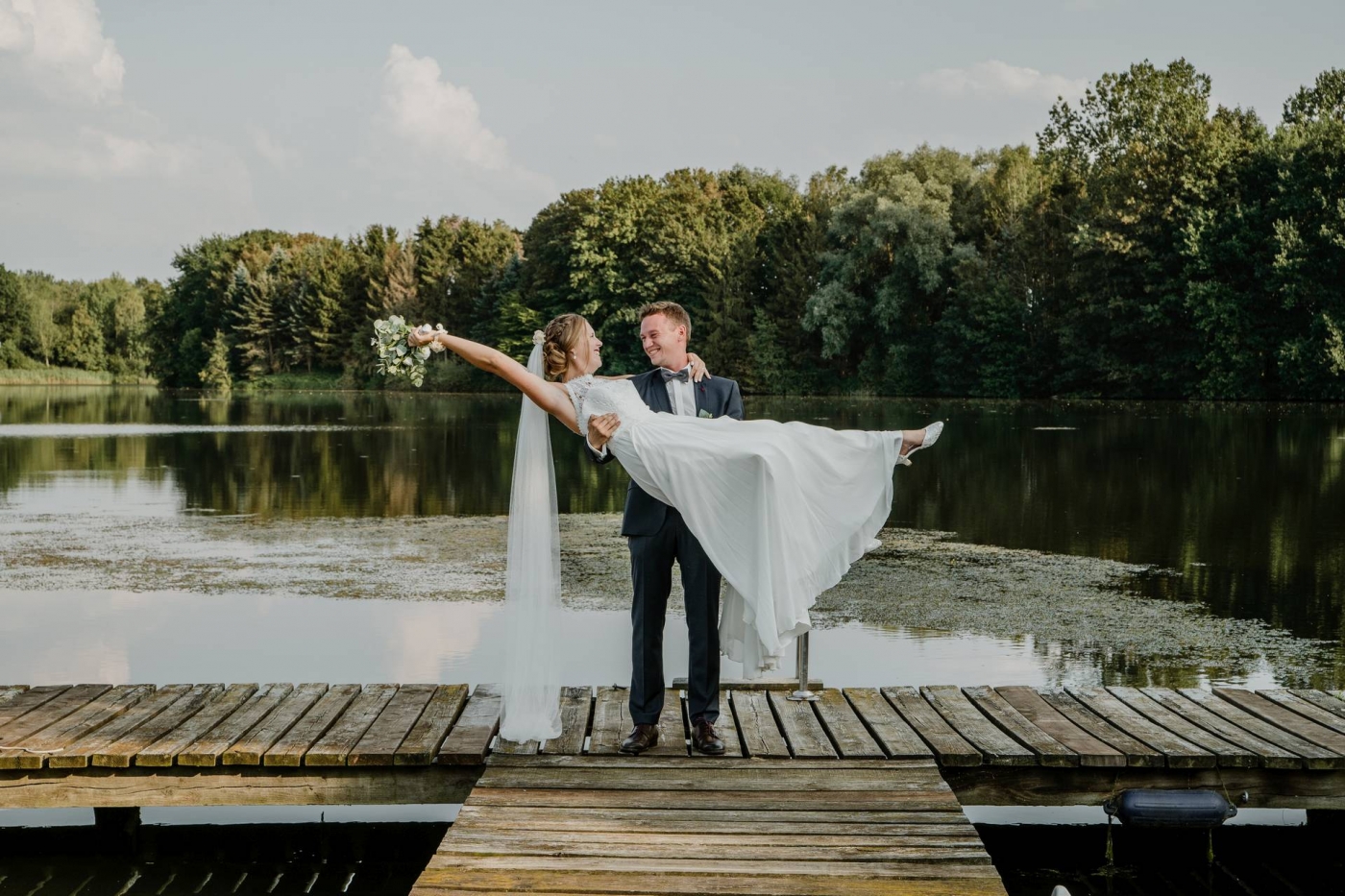 Bräutigam trägt Braut im Hochzeitskleid auf einem Steg am See, umgeben von Blumen und Bäumen.