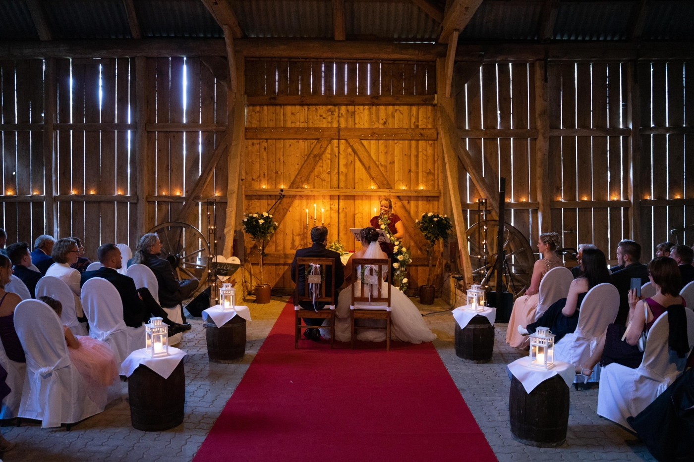 Eine Braut und ein Bräutigam sitzen bei ihrer Hochzeit in einer rustikalen Scheune, umgeben von Gästen und festlicher Dekoration.