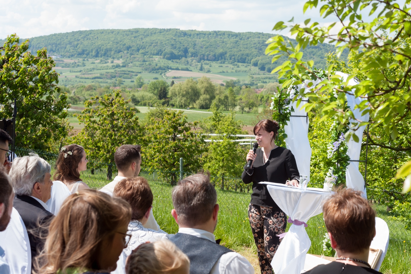 Hochzeitsfoto: Braut und Bräutigam bei Trauung im Freien, umgeben von Gästen, Hochzeitsdekoration und malerischer Landschaft.