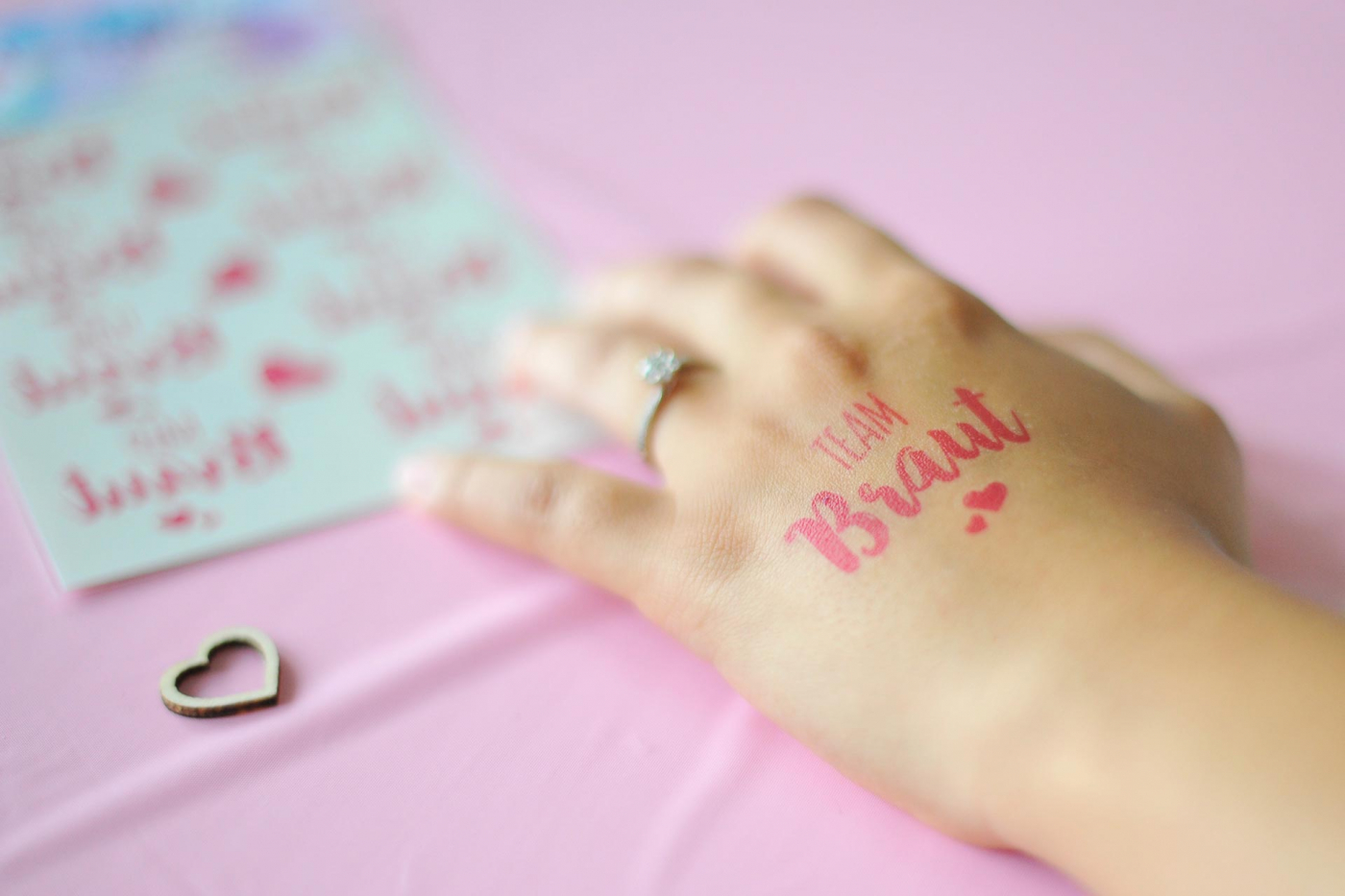 Nahaufnahme einer Hand mit "Team Braut" Tattoo und Verlobungsring, Holzdauerdekoration und Hochzeitstatoos auf rosafarbenem Tisch.