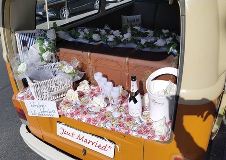 Dekorierter Retro-Bus mit Hochzeitsblumen, Champagner, und "Just Married"-Schild im Kofferraum. Optimal für Hochzeitsfotografie.