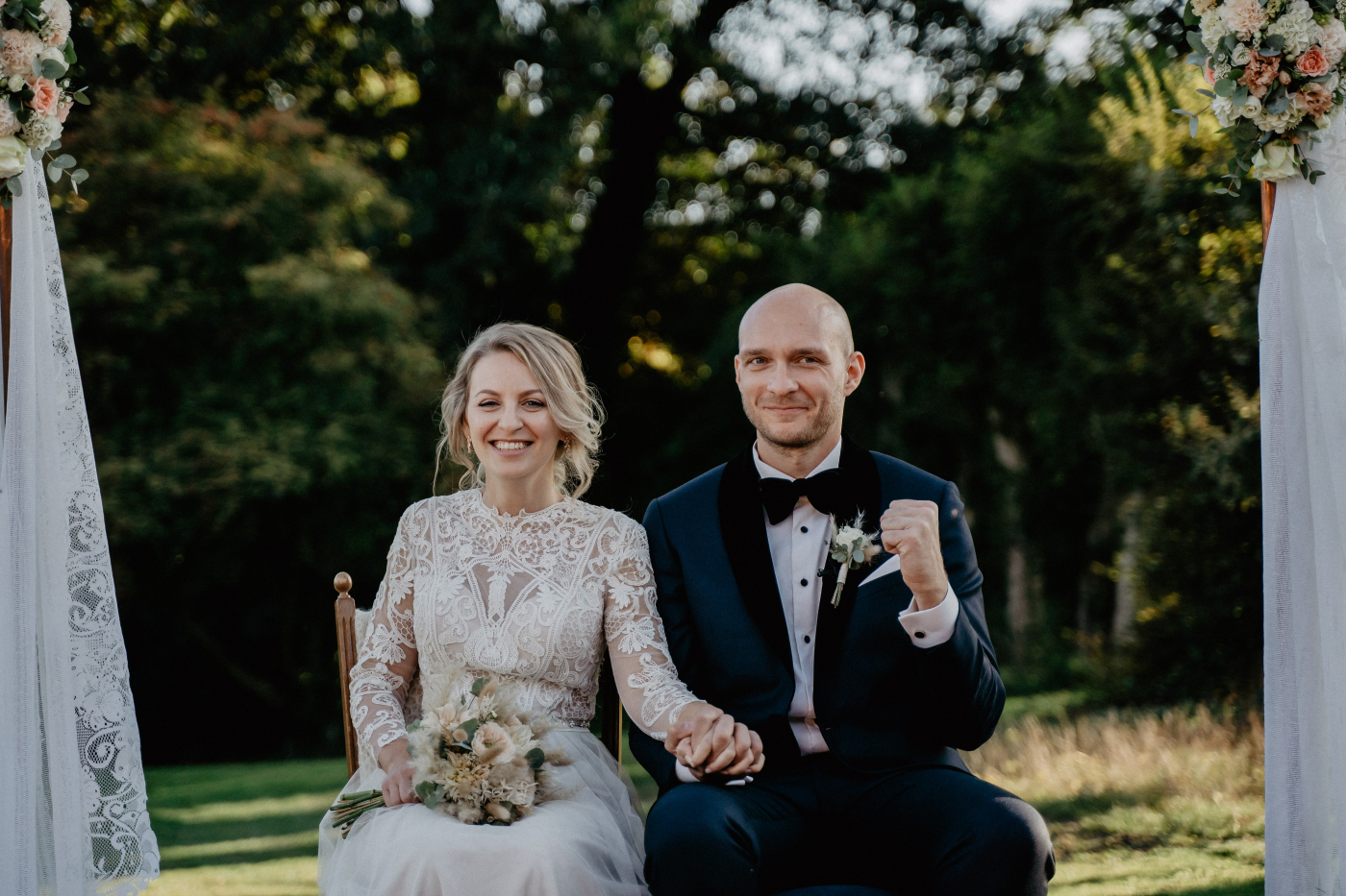 Emotionales Brautpaar unter Hochzeitsbogen, Hand in Hand, nach Hochzeitszeremonie im Garten. Bräutigam zeigt Freude.