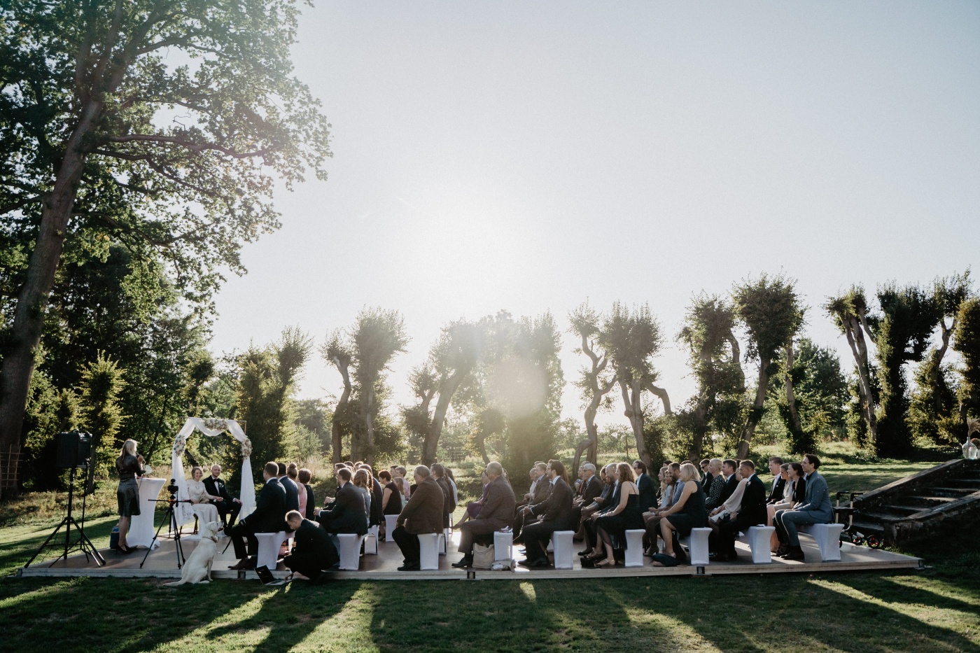 Gäste sitzen unter Bäumen auf Stühlen und warten auf die Trauung vor einem geschmückten Hochzeitsbogen.