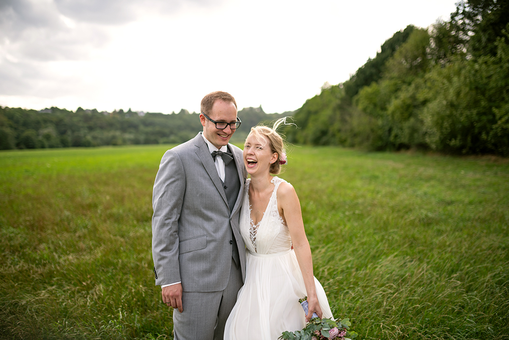 Ein glückliches Brautpaar in Hochzeitskleidung, lachend in einer grünen Wiesenlandschaft nach der Hochzeitszeremonie.