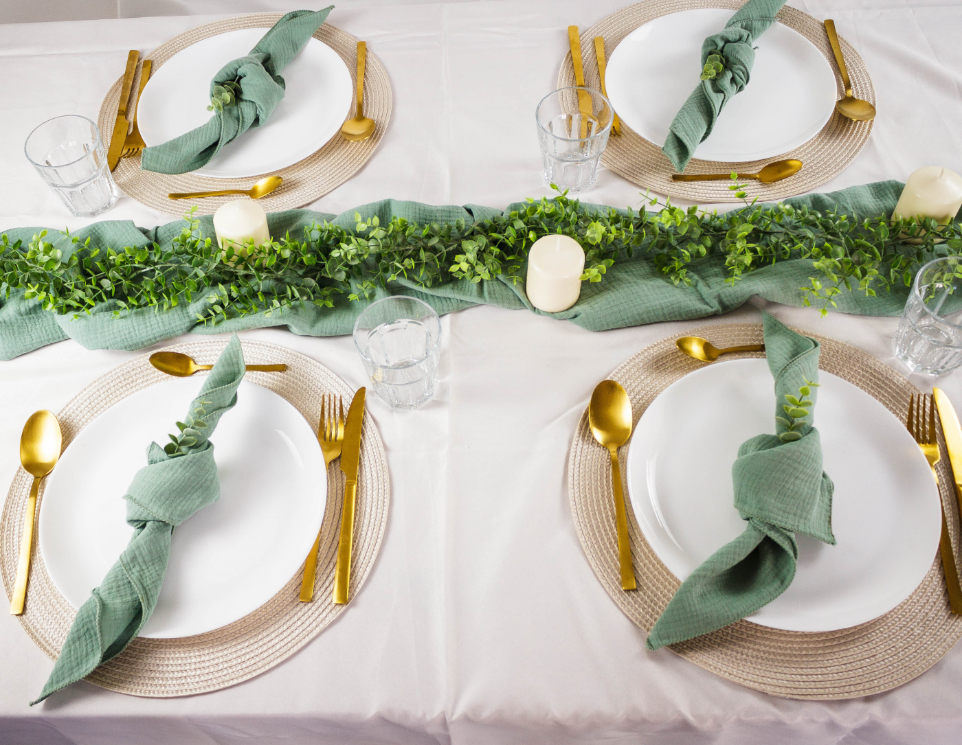 Tischgedeck mit goldenen Bestecken, grünen Servietten und blumiger Tischdekoration auf weißer Tischdecke.