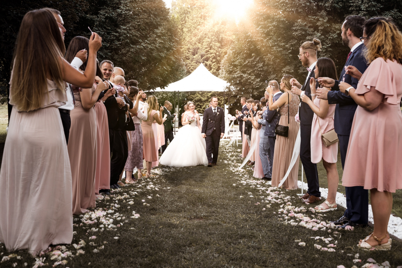 Braut und Bräutigam gehen mit Hochzeitsgesellschaft durch Blumenallee zur Zeremonie. Hochzeitskleid und Dekoration gut sichtbar