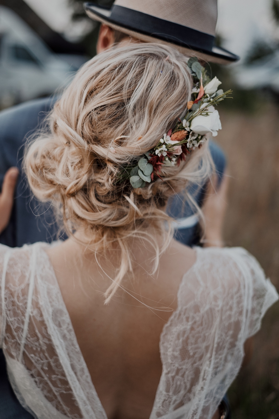 Braut mit Blumen im Haar und Spitzen-Hochzeitskleid umarmt Bräutigam; Hochzeitsfotografie im Freien.