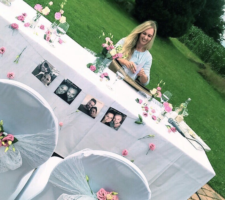 Brautjungfer sitzt an einem Hochzeitstisch im Freien, dekoriert mit Blumen, Fotos und gedeckten Stühlen.