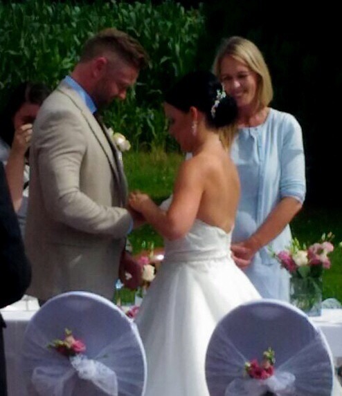 Braut steckt Bräutigam bei einer Hochzeitszeremonie im Freien den Ehering an. Hochzeitskleid, Blumen und Stuhldekoration sind sichtbar.